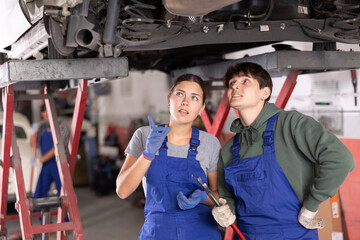 Two focused young auto mechanics, girl and guy, in blue overalls working together, inspecting car...