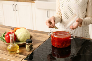 Young woman adding bay leaves to pot with delicious borscht in kitchen, closeup