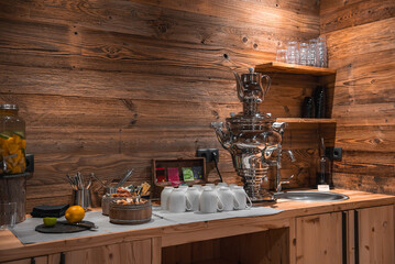 Cozy refreshment area with wooden walls, a polished samovar, teacups, tea selection, snacks, and infused water in an alpine hotel setting.