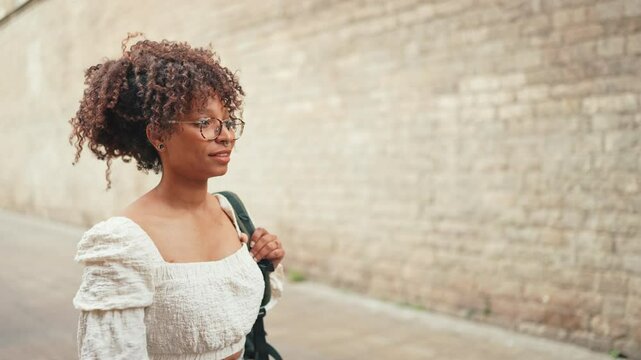 Young woman in glasses walks down the street along a brick wall. Girl goes on the street in urban background