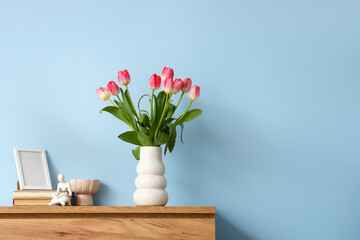 Vase with tulips for Women's Day, blank frame and books on chest of drawers near blue wall