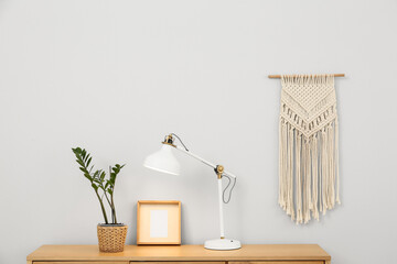 Wooden chest of drawers with lamp, houseplant and frame near light wall in room