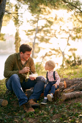 Smiling dad with lunchbox sitting near little girl on logs in forest