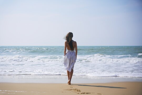 A girl in a white and blue summer dress walks barefoot along the sandy beach, heading toward the gentle sea. Her hair flows in the wind, capturing a serene and romantic seaside moment. 