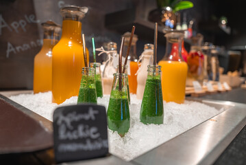 Glass bottles of green smoothies with straws and carafes of orange and yellow juices on ice, set in...