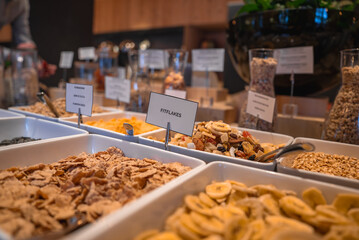 A breakfast buffet with cereals, dried fruits, and granola in white trays, labeled clearly. Glass jars and greenery decorate the elegant dining area.