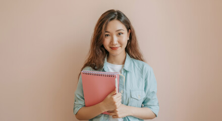 Fototapeta premium Smiling asian female teen holding notebook against pink background
