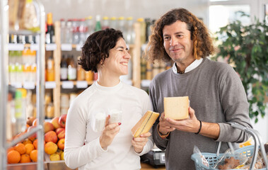 Couple choosing compare varieties some sorts of cheese, doubt and think before buying. Client choose and decide purchase cheese delicious package, inspect good.