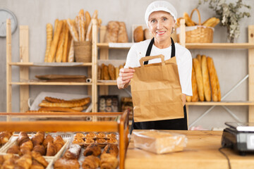 Positive mature saleswoman giving paper bag containing baked goods in bakery shop
