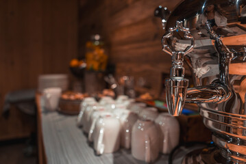 Cozy indoor scene with a metal beverage dispenser, white ceramic cups, pastries, orange juice, wooden walls, and warm lighting in the Italian Alps.