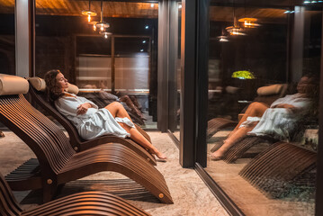 A woman in a white robe reclines on a curved wooden lounge chair near a glass window. The space features stone flooring and modern pendant lighting. © Aerial Film Studio
