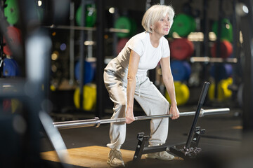 Modern motivated fit senior woman performing deadlifts with barbell during regular strength...