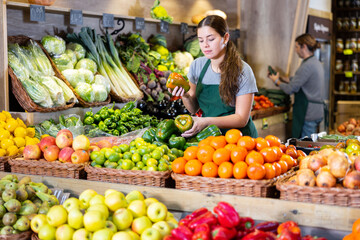 Friendly woman seller laying out ripe bell peppers on a showcase in a supermarket