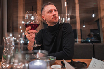 A man sits at a table in a modern restaurant with wooden interiors, holding a glass of red wine. The setting includes warm lighting and large glass windows.