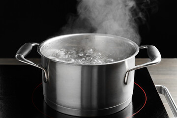 Cooking pot with boiling water and stove on wooden table against black background, closeup