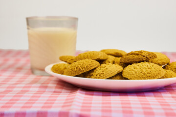 A plate of cookies sits on a pink and white checkered tablecloth next to a glass of milk. The cookies are arranged in a pyramid shape, with the top cookie being  concept of comfort and pleasure.