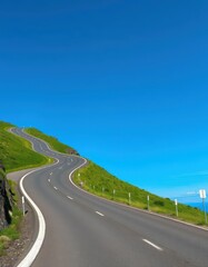 Fototapeta premium Winding asphalt road climbs a green hill against a vibrant blue sky, highway, countryside, sky