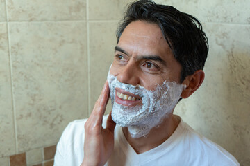Close-up of a Latino man in a white t-shirt applying shaving foam to his beard in his bathroom and...