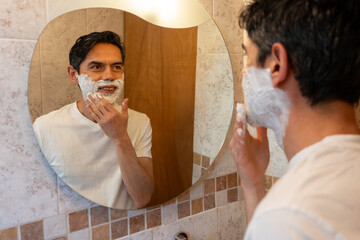Latino man looking in the mirror at how to apply shaving foam to his beard in his bathroom at home.