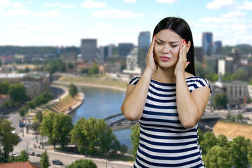 Young asian woman is suffering from headache outdoors. Urban cityscape background.