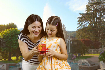 Fototapeta premium Young brunette woman and little girl open a red gift box together. Summer sunny park background.