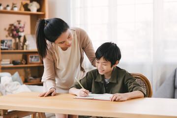 Smiling Asian mother and son bonding while studying at home desk. The room's cozy atmosphere...