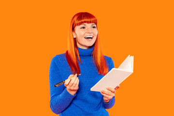 Red-haired woman smiling while holding a notebook and pen in a vibrant orange background capturing...