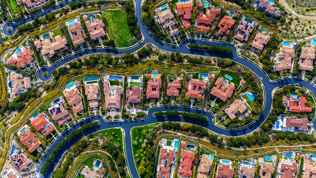 High-resolution aerial photograph showing a planned neighborhood with curved streets and luxury homes with red tile roofs and backyard swimming pools in Newport Beach, California. The community is sur