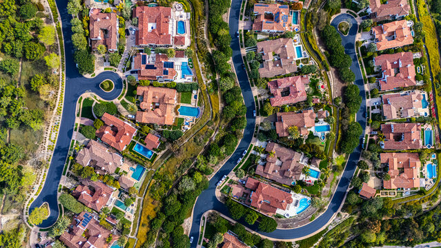 High-resolution aerial photograph showing a planned neighborhood with curved streets and luxury homes with red tile roofs and backyard swimming pools in Newport Beach, California. The community is sur