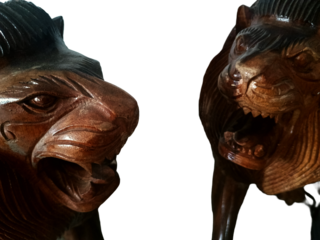 Two wooden carvings of lion heads with open mouths showcasing teeth and tongues. Intricate detailing highlights the lifelike expressions, textured manes, and masterful wood craftsmanship.