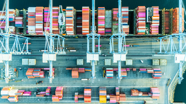 A straight-down aerial photo of shipping cranes operating over a dock filled with colorful cargo containers. The image highlights the symmetry and industrial activity of a modern shipping port.