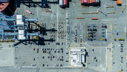 Top-down aerial view of a busy port terminal showing rows of parked cars, shipping containers, cranes, and service vehicles. The image captures the scale and organisation of a logistics and transport