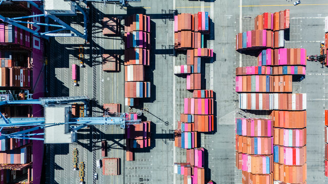 A straight-down aerial photo of shipping cranes operating over a dock filled with colorful cargo containers. The image highlights the symmetry and industrial activity of a modern shipping port.