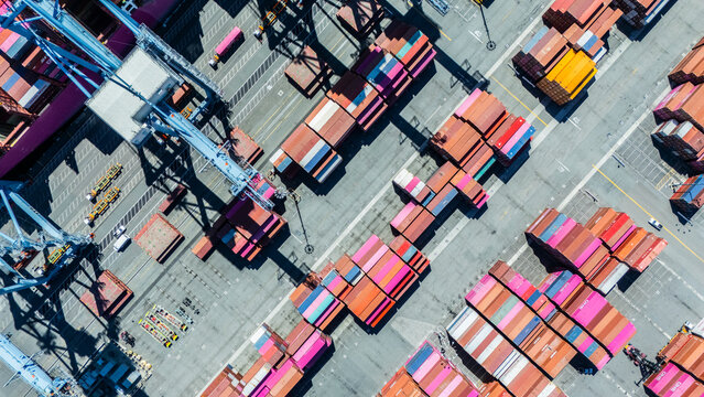 A straight-down aerial photo of shipping cranes operating over a dock filled with colorful cargo containers. The image highlights the symmetry and industrial activity of a modern shipping port.