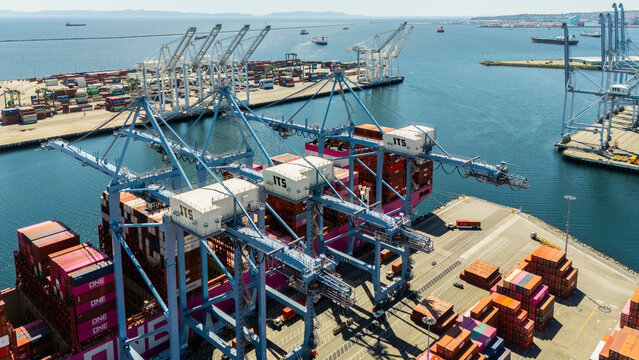 High-angle view of towering shipping cranes moving containers from a large cargo ship docked at a seaport. The deep blue ocean and colorful containers create a dynamic logistics scene.