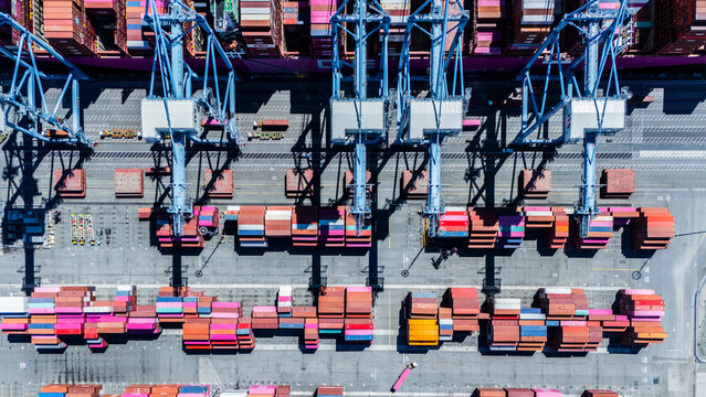 A straight-down aerial photo of shipping cranes operating over a dock filled with colorful cargo containers. The image highlights the symmetry and industrial activity of a modern shipping port.