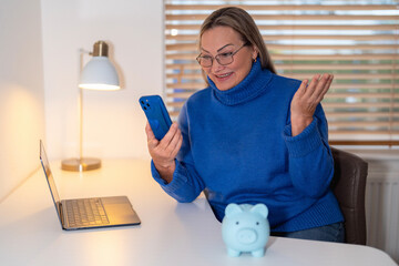 Woman in blue sweater smiles while using smartphone at desk with laptop and piggy bank during...