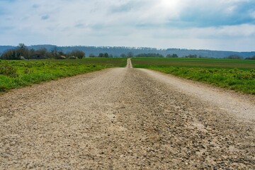 A long gravel road stretches through green fields under a partly cloudy sky. The sense of an...