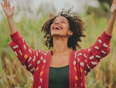 A joyful young woman with curly hair laughs ecstatically outdoors in a field of tall grass with her arms raised high to the sky feeling free and happy.