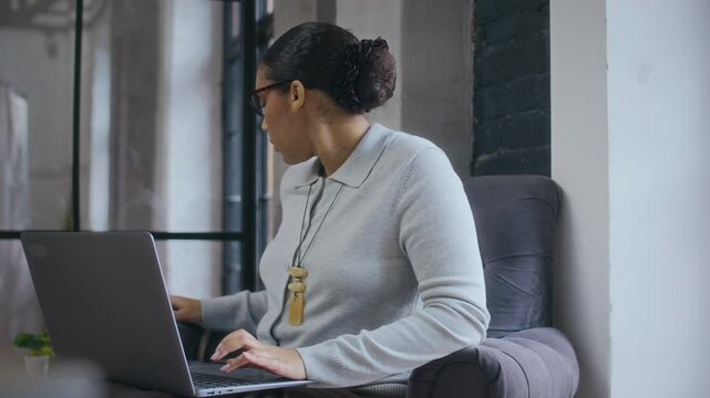 Medium shot of young African American businesswoman sitting in armchair in modern office and working on laptop, then checking messages on mobile phone and smiling