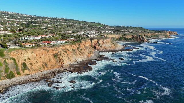 A dramatic side view of bright orange cliffs in Rancho Palos Verdes, Los Angeles, lined with luxurious homes overlooking the Pacific Ocean. Captures the unique coastal geography of California.