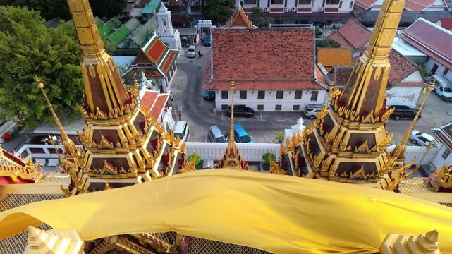 High-angle aerial view of golden temple spires and rooftops in Bangkok as a yellow sacred drapery flutters in the wind at Wat Ratchanatdaram Worawihan loha prasat in Bangkok