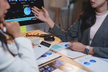 Two businesswomen collaborating on a project, analyzing financial charts and data on a computer screen while discussing key performance indicators with a calculator and reports