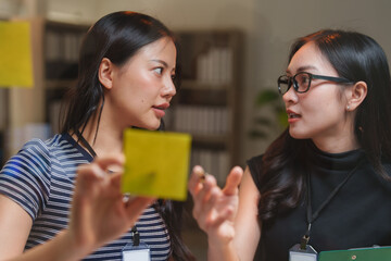 Two asian businesswomen are discussing and sharing ideas using sticky notes during a brainstorming...