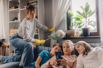Mother dusting while children using smartphone on sofa
