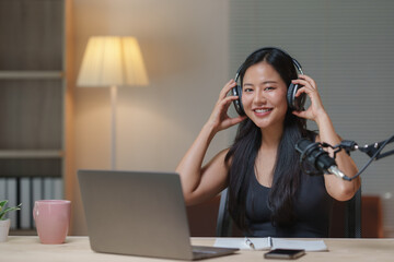 Happy podcaster wearing headphones and adjusting a microphone while recording an engaging audio podcast in a cozy home studio, surrounded by a laptop and a notebook