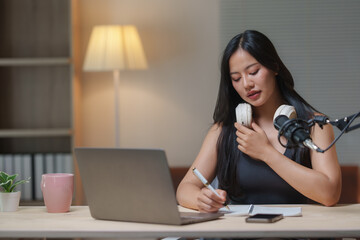 Young asian woman preparing for a podcast episode, holding headphones around her neck and taking notes while sitting at her desk with a microphone and laptop