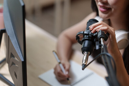 Asian female podcaster speaking into a microphone, taking notes, and recording a podcast episode in a cozy home studio filled with audio equipment and creative energy