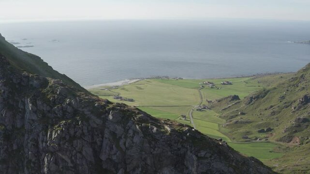 Wide aerial view from Mannen in Vestv&aring;g&oslash;y, Lofoten, Norway. Going over mountain ridge with hiking trail with green fields and the vast ocean in the background. Drone pushing in, camera tilting down.