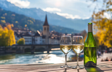 bottle of white wine and two glasses sit on table overlooking scenic river and church in picturesque town, with mountains in background. setting is serene and inviting